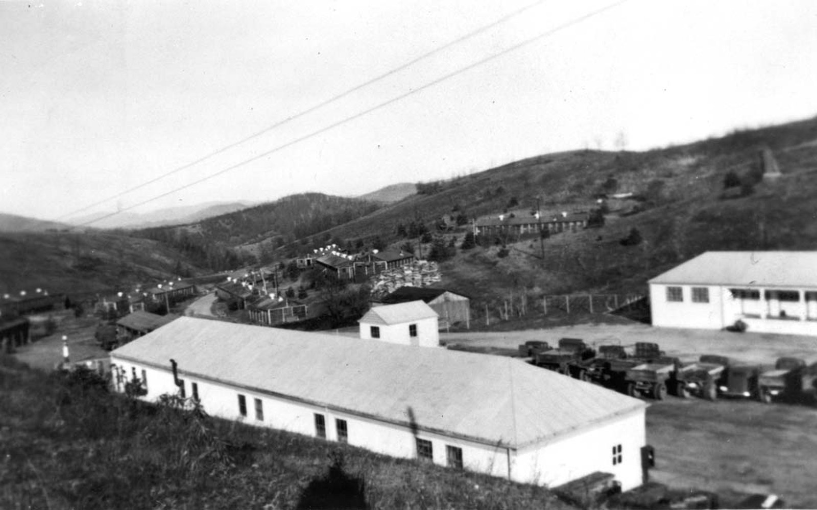Long barracks-style buildings and parked vehicles in a valley construction camp surrounded by hills