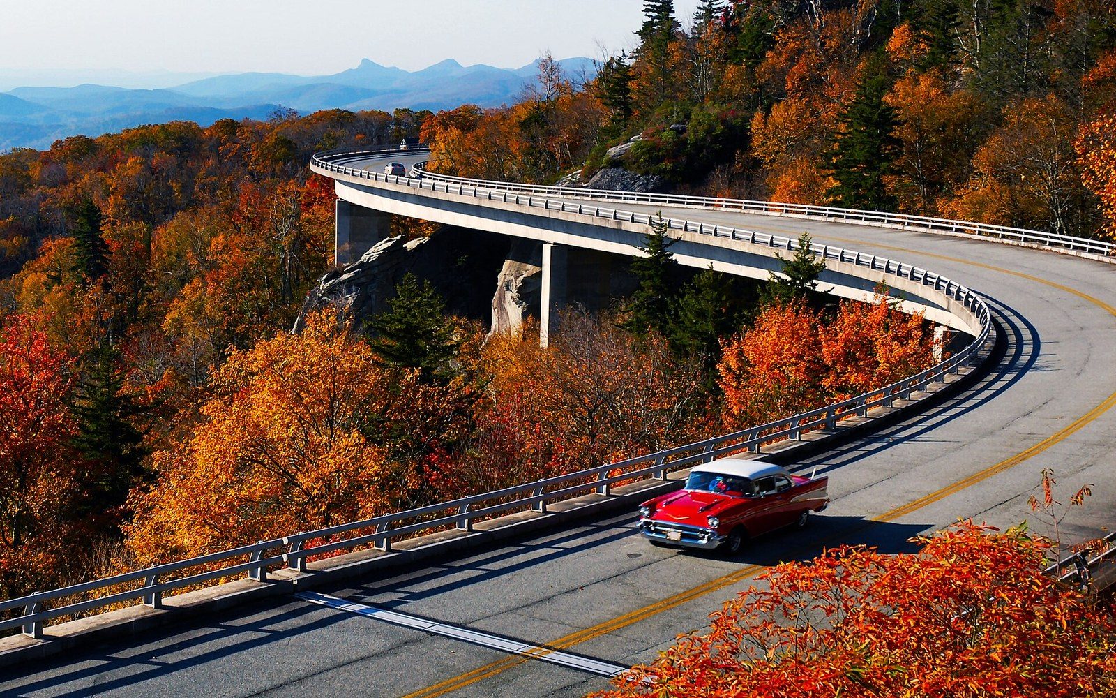 A vintage red car drives along the curving Linn Cove Viaduct amid autumn foliage