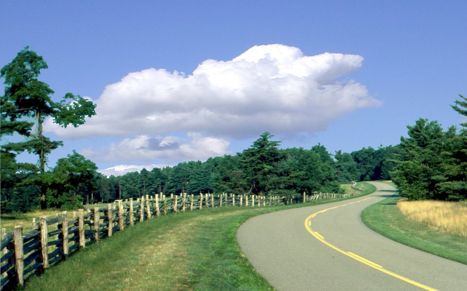 A winding two-lane parkway curves past a split-rail fence under a large puffy cloud