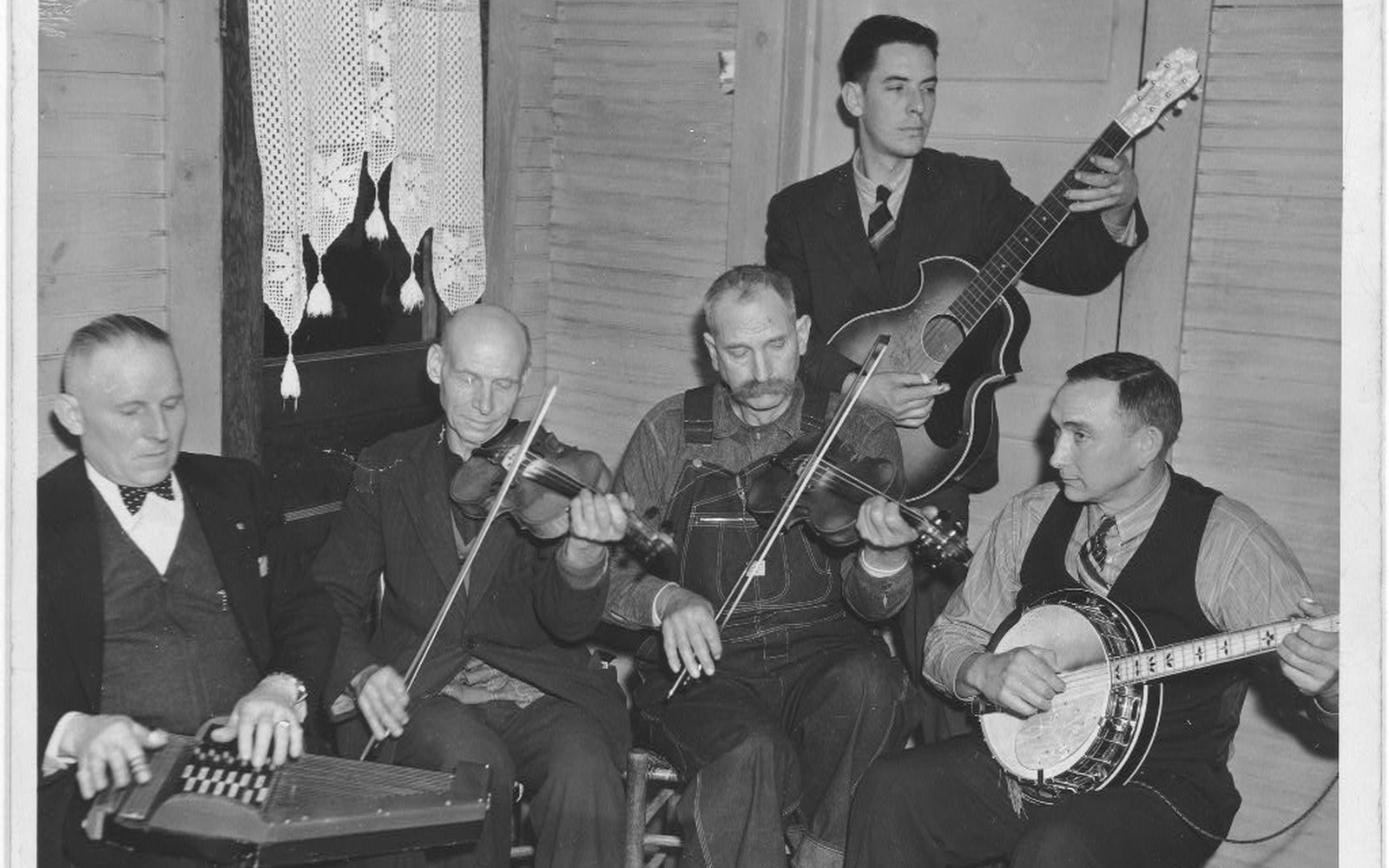 Five men play zither, two fiddles, guitar and banjo together in a front room